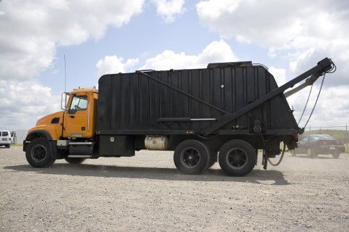A large industrial waste collection vehicle with a yellow cab and black rear compactor parked on a gravel surface under a partly cloudy sky. The vehicle features a long, sloped loading area with an attached hydraulic arm and lifting mechanism, used for hauling and disposing of bulky rubbish. The rear section is enclosed with vertical ridges, and visible stabilisers support the structure. In the background, there are other vehicles, including a white van and a silver car, parked nearby on an open lot. The scene suggests a commercial waste removal operation possibly within or near Enfield, with the vehicle ready for rubbish collection or disposal tasks, aligned with services offered by Commercial Waste Removal Enfield.