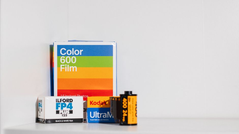 A close-up image of various photography and film consumables placed on a plain, light-colored surface against a neutral grey background. The items include a box of color film labeled 'Color 600 Film' 