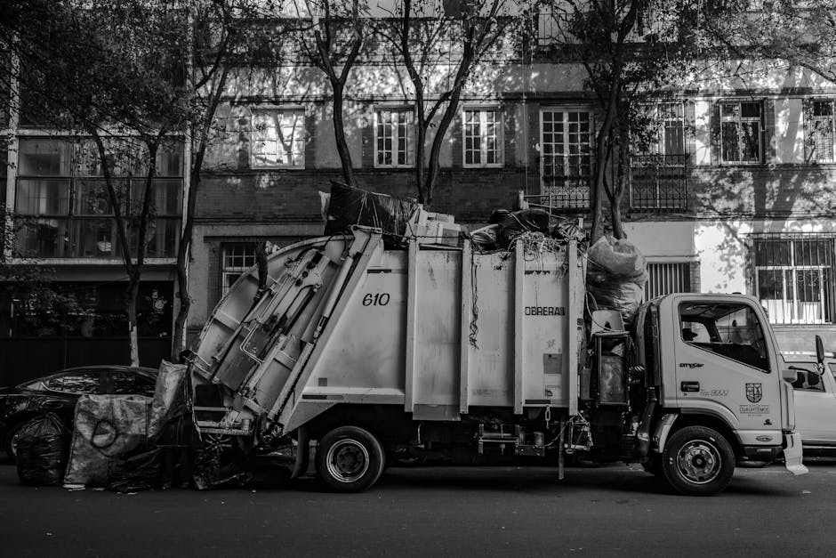 A row of four wheeled rubbish bins positioned on a cobblestone street in front of a peach-colored building with three windows featuring metal security grills. The first bin on the left is dark grey wi